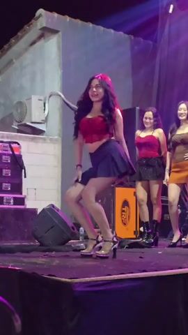 A group of young women are performing on a stage under purple and pink lights. The woman in the foreground is wearing a red crop top and a black pleated skirt with high heels. She is dancing and smiling. Behind her, two other women are also on stage, both wearing similar outfits. One is wearing black boots and the other has on heels. There are speakers and stage equipment around them. The background appears to be an outdoor setting at night, with a building and what looks like an air conditioning unit visible on the left.