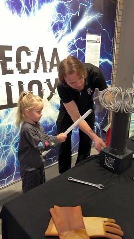 A man and a young girl stand together in front of a Tesla coil experiment, which is placed on a table covered with a black cloth. The girl, who has blonde hair in a ponytail, is holding a long, white tube that glows when the man activates the coil. The man wears a black shirt, is clean-shaven and has shoulder-length brown hair. He is wearing a glove on one hand, and is using it to help her hold the tube near the top of the Tesla coil. A wrench and a pair of rubber gloves also rest on the table. Behind them is a large sign with a blue and white graphic of lightning bolts and text that appears to be Russian. The scene takes place in a museum setting.