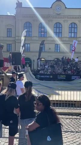 A skateboarder in a white helmet and dark clothing performs a trick on a ramp in front of a crowd. The ramp has yellow and blue accents and is set up in an urban square with a large building in the background. There are several banners visible, including ones that say "Svenska Spel" and "Karlstad." People are watching from the ramp's edge and from bleacher-style seating. The sun is shining, casting a glare across the scene.