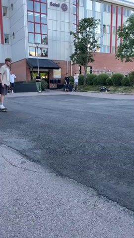 In this short video, a young man is skateboarding in an outdoor urban environment. He is wearing a light-colored, short-sleeved collared shirt, dark shorts, white socks with blue stripes, and white sneakers. He is actively engaged in skateboarding, performing tricks.

The scene appears to be a paved area, possibly a parking lot or a designated skateboarding area, with a ramp or obstacle in the middle. The ramp has "PLAZA" written on it in large letters, along with graffiti tags. The background shows buildings, trees, parked cars, and other people observing or participating in the activity. One building in the background has a sign that reads "Suttonss Solle".

The time of day seems to be late afternoon or early evening, as indicated by the lighting and the sky. The weather appears to be pleasant and sunny, with a clear sky. The overall atmosphere seems casual and recreational, with people enjoying themselves. The skateboarder's actions suggest a focus on practice and skill development in skateboarding.