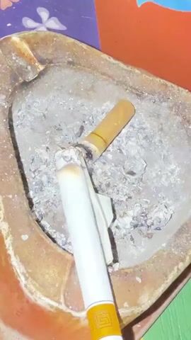 A close-up, overhead shot focuses on an ashtray filled with cigarette ash and two cigarette butts. One cigarette butt, longer and mostly burned, rests at an angle in the ashtray. A second, mostly intact cigarette, lies across the ash. The cigarette is white with a yellow band and logo near the filter. The ash is gray and appears to be from several extinguished cigarettes. The ashtray is made of a dark, possibly ceramic material with visible rust or staining along the rim. In the background, a colorful, patterned surface is partially visible. The lighting is dim and appears to be artificial.