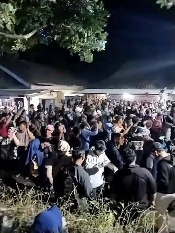 The video captures a large gathering of people outdoors at night. The crowd is densely packed, with many individuals raising their hands in the air, suggesting they are engaged in a celebratory or energetic activity, possibly a concert or a festival. A stage with lighting is visible in the background, further supporting the idea of a performance. The environment appears to be somewhat unpaved, with foliage in the foreground, indicating an outdoor, possibly informal, venue. The overall mood appears to be lively and communal.