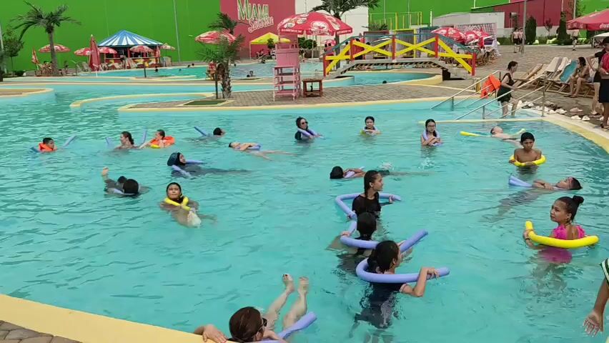 This is a medium shot of a crowded swimming pool at a water park on a bright day. The pool is blue with yellow edging and has several shallow areas with colorful umbrellas and palm trees. Many people, mostly children and young adults, are in the water, some with floatation devices like inflatable rings and pool noodles. Some are swimming, while others are playing or relaxing. In the background, there's a red building with a sign that says "Neptun," a lifeguard stand, lounge chairs, and a wooden bridge over a section of the pool. The overall atmosphere appears to be one of recreation and enjoyment.
