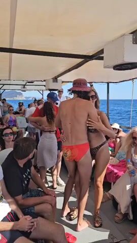 The video shows a crowded outdoor boat party. In the foreground, a couple dressed in swimwear is embracing. The man is wearing red swim trunks and a hat, while the woman is in a leopard-print bikini. They are standing between other partygoers. The boat is covered by a white canopy, and the ocean is visible in the background, with a coastline in the distance. Other people are seated and standing around the boat, some interacting with each other, while others appear to be relaxing. The overall atmosphere seems festive.