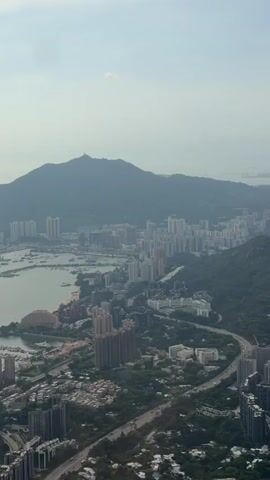 This is a vertical aerial drone shot of a dense city nestled beside a large body of water and a prominent mountain in the background. The city is characterized by numerous tall, modern buildings, many of which appear to be residential high-rises. A harbor or bay area is visible, with many boats and yachts docked. A highway curves through the city, with some traffic visible. Lush green trees and vegetation are scattered throughout the urban landscape and cover the slopes of the surrounding hills. The sky is hazy with a few clouds, suggesting an overcast or humid day. The overall impression is one of a sprawling metropolis integrated with its natural surroundings.