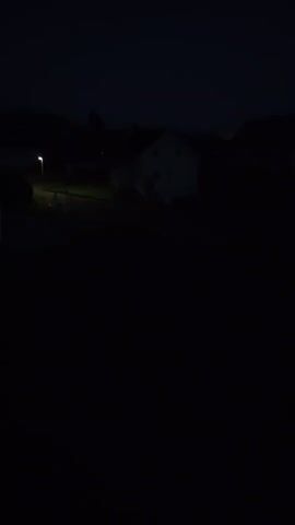 The video shows a dark, possibly rural, nighttime scene.  A house is visible in the distance, vaguely illuminated. A single street lamp casts a glow across a yard.  Mountains are silhouetted in the background against a dark sky.  Nothing else is discernible in the dark.