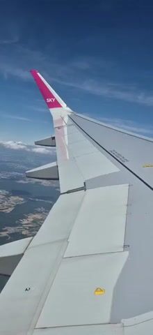 An airplane wing is visible in the foreground, with the tip of the wing featuring a pink stripe and the word "SKY" printed in white letters. Below the wing, a landscape of trees and buildings can be seen from high above. The sky is a bright blue with scattered white clouds. The wing has several mechanical components, including flaps and ailerons, and warning labels are visible on its surface. The camera is positioned to show the wing from a perspective slightly above and to the side, looking towards the horizon.