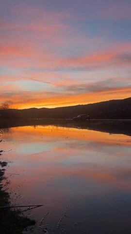 The video shows a body of water reflecting a colorful sunset. The sky is filled with orange, pink, and blue hues. In the background, there are silhouetted mountains and trees. A vehicle is seen parked near the water. The foreground shows a muddy shoreline with grass and some debris. The water is mostly still, creating a clear reflection of the sky.
