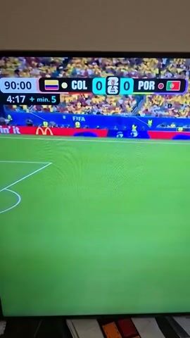The video shows a close-up of a television screen displaying a soccer match. The scoreboard at the top of the screen indicates that the match is between Colombia (COL) and Portugal (POR), with the score currently 0-0 in the 90th minute, with 4 minutes and 19 seconds of stoppage time remaining, plus an additional 5 minutes. The game is being played on a green grass field with white markings. Several soccer players in red and yellow uniforms are visible on the field. In the background, a stadium filled with spectators in yellow shirts can be seen. A banner with the McDonald's logo and the text "I'm lovin' it" is displayed below the scoreboard. The lower part of the image shows a dark surface with some papers and a small white fan.