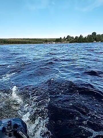 The video shows a body of water with waves. There is a wake from a boat in the foreground. In the distance, there is a shoreline with trees and buildings. The sky is blue with some clouds. The video appears to be taken from a moving boat.