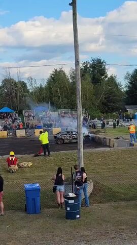 The video shows an outdoor event, possibly a demolition derby or similar spectacle. In the background, a damaged car is smoking heavily in the center of a dirt arena, surrounded by concrete barriers. Bleachers filled with spectators are visible behind the arena. Several people are on the dirt area. One person in a yellow shirt is seen spraying something from a red canister towards the smoking car. Another person in a yellow shirt stands nearby. A person in a red helmet and yellow vest is kneeling on the ground. In the foreground, four individuals are standing behind a chain-link fence. Two blue trash bins and a dark barrel are also in the foreground. Trees and a cloudy blue sky form the background. A wooden utility pole runs vertically through the center of the frame.