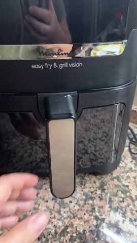 A hand is shown pulling out the drawer of a Moulinex air fryer. Inside the drawer, two golden-brown pastries are revealed, sitting on parchment paper. The pastries appear to be filled and have a lattice-like pattern on top. The air fryer is black with a chrome accent that says "Moulinex easy fry & grill vision". The scene is well-lit, and the background appears to be a kitchen countertop.