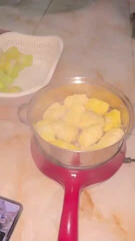A stovetop steamer with a red base and a silver pot containing multiple food items, likely cabbage rolls or dumplings, is shown. Steam is visible rising from the pot. To the left, a white plastic colander holds chopped green vegetables. A smartphone screen is partially visible in the bottom left corner. The steamer is placed on a light-colored tiled surface. The scene appears to be indoors, possibly a kitchen. The video is a short, static shot with no discernible action or interaction.