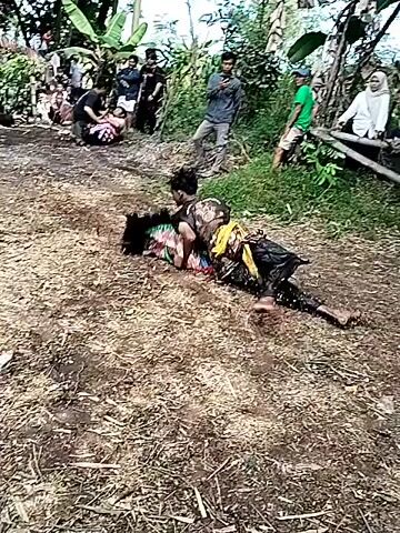 In an outdoor setting with a crowd of onlookers gathered around, two individuals are engaged in a physical struggle on the ground. The ground appears to be covered with leaves and dirt. The individuals are dressed in what appears to be traditional clothing, with one predominantly in black and the other wearing a yellow sash. They wrestle, rolling around on the ground. After a period of struggle, one of the individuals stands up and walks away, leaving the other prone on the ground. Another person approaches the fallen individual and kneels beside them. The crowd remains watching. The scene is surrounded by trees and vegetation.