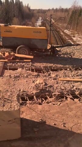 The video shows a construction site where a concrete bridge is being demolished. In the distance, a railway track stretches across the landscape, lined with trees and patches of snow. A large, red jackhammer is seen breaking up the concrete. The area is filled with debris and exposed rebar. The weather appears sunny and bright.