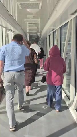 The video begins with a shot from behind a group of people walking down a brightly lit enclosed walkway with large windows on the right. The walkway appears to be an airport jet bridge or a similar transit area. The people are walking away from the camera. The first person is a man wearing a light blue short-sleeved shirt and gray pants, carrying a brown bag. Behind him is a woman in a dark brown dress. Next to her is another person wearing a burgundy hooded sweatshirt and loose-fitting blue pants. Further ahead, another person in a white shirt is visible. The walkway has a reflective ceiling with fluorescent lights and a neutral-colored floor. The windows reveal a modern building with glass facades and a bright sky. As the video progresses, the camera pans slightly to the right, offering a view through the windows of the exterior. We see a train or tram station with sleek, red and white vehicles parked at platforms. There are also signs, stairs, and a yellow vehicle that looks like a small forklift or utility vehicle on a roadway. The architecture outside is contemporary, with glass and metal structures. The camera continues to move, showing more of the station area and a street with a few cars. The overall impression is one of a modern transportation hub.
