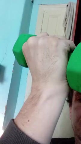 Here's a description of the video:

A man is holding two green dumbbells and doing bicep curls. He is close to the camera, and his face is partially visible. The background includes a blue wall and a white door.