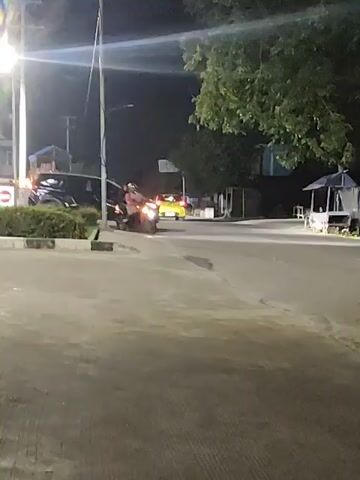At night, a black SUV is parked and turned on, with its headlights illuminated. A motorcycle with two people on it is approaching the SUV. The motorcycle is illuminated by its own headlights, which are also on. The scene is an asphalt road with a sidewalk and some trees in the background. There is also a small stand on the right side of the road.
