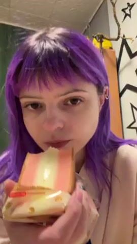 A woman with purple hair is eating a piece of cake. The cake has three layers, two pink and one white. The woman takes a large bite. She has earrings in both ears. There is art on the wall behind her and a textured ceiling. The cake is partially covered in plastic wrap. She holds the cake in both hands while eating.