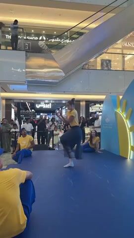 A group of performers are on a blue stage in a mall. A woman in a yellow shirt and dark skirt is dancing center stage. Two people in yellow shirts and blue pants are sitting on the stage. A crowd watches them from a distance. The mall has escalators and shops in the background.