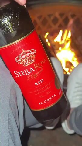 The video shows a first-person perspective of someone holding a red bottle with a label that says "STELLA ROSA" and "ROSE" in white text. The bottle is positioned in the foreground, slightly out of focus, obscuring a portion of the scene. In the background, a bonfire is burning brightly, with logs crackling and flames dancing. In front of the bonfire, a pair of white sneakers is visible, suggesting the person is sitting with their feet extended towards the fire. The environment appears to be outdoors, possibly a backyard or campsite, given the presence of the fire pit. The lighting is dim, with the fire providing the primary illumination.