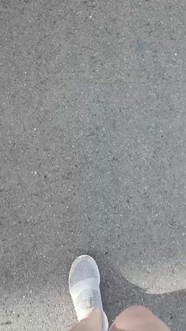 This video shows a first-person perspective of someone walking along a paved road in a rural or suburban area. The road surface is asphalt with visible aggregate. The person is wearing a gray athletic shoe with a white sole and red shorts. The scene suggests an outdoor environment with daylight, indicated by the shadows and the overall brightness of the footage.

In the background, there are houses or buildings, some with fences and vegetation. Several cars are parked along the roadside, including a silver hatchback, a white SUV, and part of another silver vehicle. Utility poles and wires are also visible, suggesting infrastructure typical of a residential area. The weather appears to be clear or partly cloudy, with a bright sky.

The activity taking place is simply walking. There are no specific events or interactions depicted. The mood is neutral and observational, focusing on the act of walking and the surrounding environment. The location cues, such as the style of houses and cars, along with the provided context of Miasskoye, Russia, help to establish the setting. No visible text is present in the video.