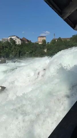 This video captures a wide shot of the Rhine Falls in Nohl, Switzerland, on a clear, sunny day. The powerful waterfall dominates the foreground with its immense volume of white, churning water cascading over rocks. In the background, lush green trees line the riverbanks, interspersed with buildings and structures of varying heights, suggesting a town or city nearby. A construction crane is visible, indicating ongoing development in the area. The bright sunlight and clear blue sky suggest pleasant weather conditions. The dynamic and forceful nature of the waterfall evokes a sense of awe and the raw power of nature. No visible text is present in the video.