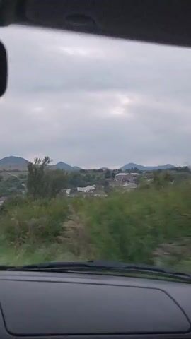 The video is taken from inside a vehicle, looking out through the windshield. The view is of a rural landscape with rolling hills covered in green vegetation and scattered houses. The sky is overcast with a light gray cloud cover. There are trees and bushes visible in the foreground, and a fence runs along the side of the road. The perspective suggests the vehicle is moving, as the landscape appears to be shifting slightly. The overall impression is one of a calm, somewhat subdued natural environment.