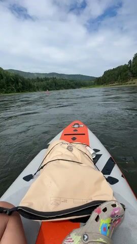 The video shows a first-person perspective from a stand-up paddleboard on a wide river. The river flows through a lush, green valley with forested hills on either side. The sky is partly cloudy, suggesting a pleasant day. In the distance, another paddleboarder can be seen. The paddleboard itself is orange and white with black accents. A beige dry bag is secured to the front of the board. A person's leg, wearing a colorful Croc shoe, is visible in the foreground. The setting appears to be Усть-Мана, Russia, indicated by the landscape of rolling hills and a substantial river. The overall mood is one of peaceful outdoor recreation.