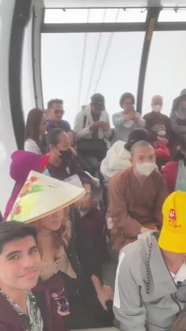The video shows a diverse group of people sitting inside a cable car. The camera is positioned low, focusing on the faces of several individuals in the foreground, including a man with dark curly hair wearing a maroon hoodie and a woman wearing a conical hat. In the background, more people are visible, some looking at their phones, others talking or observing the surroundings. Several individuals are wearing masks, and one person is dressed in a monastic robe. The interior of the cable car is visible, with the white ceiling and curved supports. The overall atmosphere appears to be one of travel or tourism.