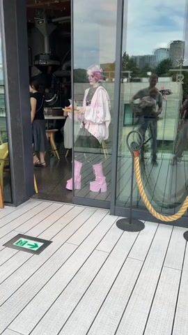 A young person with brightly colored rainbow hair and pink platform boots exits a building holding a tray with a drink. They are wearing a pink hooded jacket over a black lace top and a grey pleated skirt. The person is walking on a wooden deck. In the background, through a glass door, another person is visible, and there are buildings outside. A green exit sign is on the deck floor.