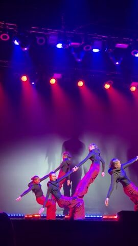 Five female dancers perform on a stage illuminated by overhead blue and red spotlights. They wear black long-sleeved shirts and baggy red pants. A large, blurry, dark silhouette of a human figure is projected onto the back wall. Initially, the dancers are in a line, holding a connected pose with extended arms and leaning bodies, some with one leg raised. They then transition into a dynamic, synchronized routine, crouching and moving their arms and legs in a fluid, hip-hop-influenced style. The silhouette remains visible in the background.