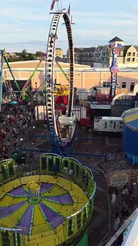 The video shows an aerial view of a busy amusement park or carnival. In the foreground, a circular ride with purple and yellow sections is visible, with several people standing on it. Behind this, a large, tall amusement ride with a circular track that goes upside down is in motion, with people visible in the cars. A Ferris wheel can be seen to the left of this ride. The park appears to be crowded with many people walking around. In the background, buildings and a cloudy sky can be seen. The overall scene depicts a lively and active atmosphere.