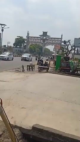 A street scene with an ornate archway in the background. Several motorcycles and a white car are visible on the road. In the foreground, there are two small tables with chairs, and a person sitting on one of the chairs. A green barrier is also present on the right side of the frame, obscuring some objects behind it. The sky is overcast.