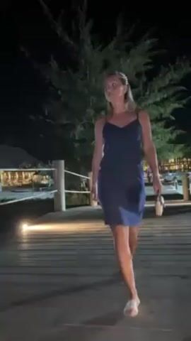 A woman is walking towards the camera at night on a boardwalk. She is wearing a navy blue, short, sleeveless dress with spaghetti straps and white sandals. She is carrying a small, light-colored purse. She has a pearl headband on her blond hair. The boardwalk is lit with small lights. Behind her are trees and buildings with lights.