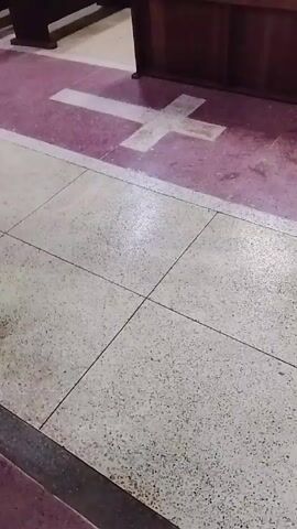 The video shows a section of a church floor with light-colored square tiles. A purple section with a white cross design is also visible near a wooden pew.  Singing is heard in the background.