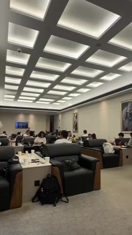 This video captures an indoor scene, likely a lounge or waiting area. The space is illuminated by a grid of recessed ceiling lights, creating a modern and bright atmosphere. Comfortable black leather armchairs are arranged in small clusters around low tables. The walls are a neutral tone, adorned with framed artwork. Several individuals are seated in the armchairs, engaged in various activities such as looking at phones, conversing, or working on laptops. Some tables have drinks and small items on them. A backpack is visible on the floor next to one of the chairs in the foreground. The overall impression is one of a calm and contemporary gathering space.