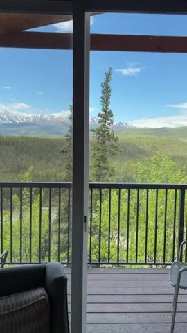 A slow pan captures a view from inside a room looking out onto a balcony and a vast natural landscape. The interior features a dark grey sofa with a patterned pillow in the foreground, partially obscuring a dark grey curtain. A large glass sliding door leads to an outdoor balcony with a grey railing. Beyond the balcony, a river winds through a valley covered in dense green trees. In the distance, snow-capped mountains rise against a bright blue sky dotted with white clouds. The video pans further to reveal a floor lamp with a white shade on the right side of the frame, positioned near a wooden wall. Another section shows a broader view of the river and a road below, framed by trees and mountains under a partly cloudy sky. The camera movement is smooth and deliberate, showcasing the expansive scenery and the transition from the interior to the exterior view.