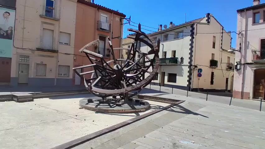 This media file captures a vibrant outdoor scene in Salt, Spain, under a bright, sunny sky, suggesting midday. The focal point is a large, intricate metal sculpture resembling a celestial globe or an astrolabe. Its weathered, dark metal construction forms a complex network of interconnected rings and spokes, some of which appear to have lettering inscribed on them. The sculpture is positioned in a paved public square, likely a plaza, with several buildings framing the background.

These buildings are multi-story structures with distinct architectural styles. On the left, a building features a light-colored facade with windows and balconies. To its right, a taller building displays a warm, orange-toned facade, also with balconies. Further in the background, a predominantly white building with dark trim and windows stands out. The presence of balconies and windows suggests residential or commercial use.

The ground is paved with concrete or similar material, showing some wear and tear. There are dark metal railings or borders defining areas of the plaza, and the sculpture itself rests on a circular base. The lighting indicates a clear day with strong sunlight casting distinct shadows.

While there are no people actively engaged in the scene, a large mural or artwork featuring figures is visible on the far left building, adding a touch of artistic expression to the environment. The overall atmosphere is one of a quiet, sun-drenched public space, possibly a historical or cultural hub given the prominent sculpture. The inscription on the sculpture, though partially obscured, appears to include the words "A SOLEDAD" and "LLAMA," which may offer clues to its artistic intent or historical significance. The weather is clearly fair and the time of day is likely around noon or early afternoon due to the bright sunlight and sharp shadows.