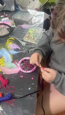 The video shows a close-up view of a person's hands as they create a dreamcatcher. The person is sitting at a table covered with craft supplies, including beads, feathers, and string. The dreamcatcher is partially completed, with a pink ring and a web of pink string. The person is adding beads to the dreamcatcher, using a needle and thread. The scene is brightly lit, and the table is messy, with paint splatters and other debris. In the background, there are several storage containers filled with beads and other craft supplies. The person's hands are adorned with nail polish. The overall impression is one of a creative and engaging activity.