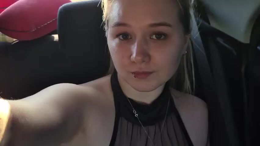 A young woman with blonde hair and light-colored eyes is shown taking a selfie inside a vehicle. She is wearing a black halter-style top and a silver necklace. There is a red pillow in the background. Sunlight streams into the car, creating a lens flare effect. The woman's facial expression is neutral, and she is looking at the camera.