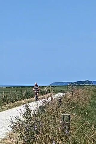 The video shows a cyclist riding on a path toward a large medieval abbey built on a rocky island. The abbey is surrounded by a flat, green landscape and a clear blue sky. The camera pans from the cyclist to the abbey, then focuses on the abbey and the surrounding landscape. There are a few fence posts and some tall grass in the foreground.