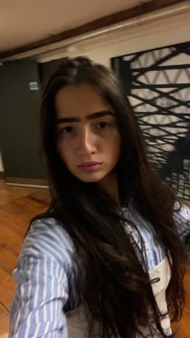 A young woman with long, dark hair stands in a hallway. She is wearing a blue and white striped shirt. The hallway has wooden floors, white brick walls, and a door with the number 502 on it. A fire extinguisher is visible on the wall. The woman is looking at the camera and making a kissing face. The camera movement is somewhat shaky, suggesting it is hand-held.