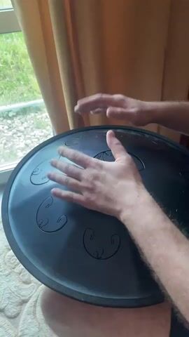 A person is playing a handpan, a steel percussion instrument. The handpan is black and circular, with several indentations on its surface that are struck to produce different tones. The person's hands are visible, moving rhythmically across the handpan. The room has a window with a view of greenery outside. A beige curtain hangs beside the window. The handpan rests on the person's lap, and the carpet is visible below.