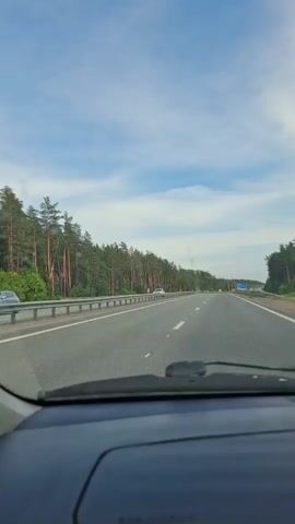 The video shows a view from inside a car driving on a highway. The car is moving forward on a paved road with two lanes in each direction, separated by a white line. A guardrail runs along the left side of the road, bordering a dense forest of tall, thin pine trees. The sky above is a mix of blue with scattered white clouds. Other vehicles, including cars and a white SUV, are visible on the road ahead and in the distance. A blue sign with white lettering is visible on the right side of the highway. The dashboard of the car is visible in the foreground. The scene appears to be during daylight hours.