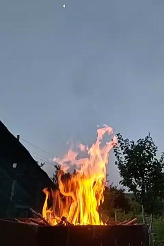 A fire burns in a metal fire pit outdoors at dusk. The flames are tall and bright orange and yellow, flickering upwards towards a pale grey sky. A faint crescent moon is visible in the sky. To the left of the fire pit is a dark, sloped roof structure covered with black netting. Behind the fire pit, trees and foliage are visible against the darkening sky. The scene is viewed from a low angle, looking up at the fire.
