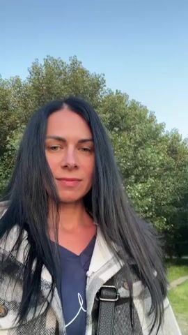 A woman with long black hair, wearing a plaid jacket over a dark blue shirt, is walking outdoors. She is holding a phone or camera in front of her, recording herself. The background shows a body of water, a grassy area, and trees. The sky is clear and appears to be either early morning or late evening, with the sun setting and casting a warm glow over the scene. The woman's face is visible, and she is looking directly at the camera.