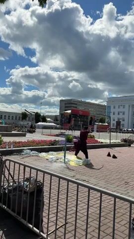 A person dressed in a burgundy outfit with a pointed hat is performing in a public square. They are holding a stick and appear to be interacting with something on the ground, possibly an object or a performance prop. Around the performer, there are scattered plastic bottles and a yellow mat. The ground is paved with red bricks. In the background, there are buildings, tents, and a large screen displaying graphics with text that appears to be in Russian. The sky is filled with white, puffy clouds, suggesting a partly cloudy day. The scene is set in a city square, possibly during an outdoor event or festival. There is a metal fence in the foreground, with a trash bag behind it. The lighting suggests it is daytime.