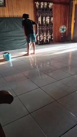 A person is mopping the floor in a room with wooden walls and tiled flooring. The person is wearing a dark t-shirt and blue shorts. A blue bucket sits on the floor next to them. A fan is visible in the background. Sunlight is streaming into the room, reflecting off the wet floor.