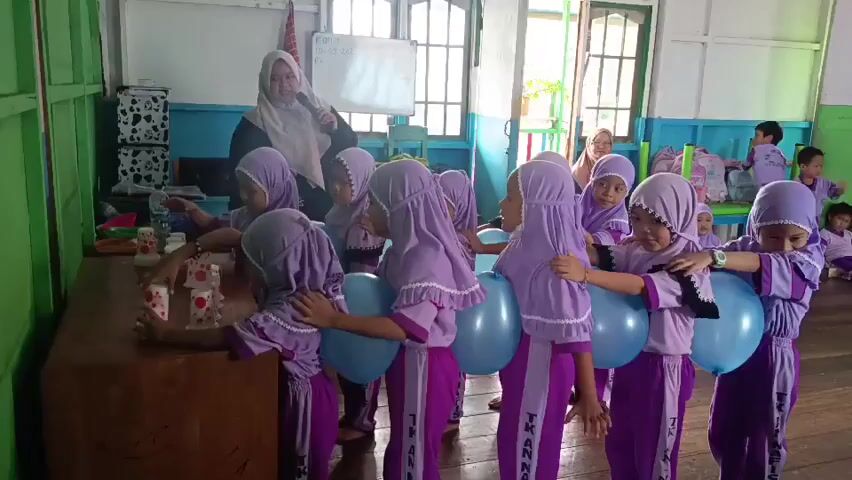 A group of young children wearing purple hijabs and matching outfits are participating in a game in a classroom. The children are arranged in a line, with some holding balloons between their legs, and others stacking cups on a table. An adult female teacher in a beige hijab stands behind them, seemingly giving instructions. The classroom appears to be a simple, somewhat rustic space with wooden floors and walls painted in different colors. There are windows in the background, suggesting natural light. Other children and adults are visible in the background. The overall atmosphere appears to be one of a playful educational activity.